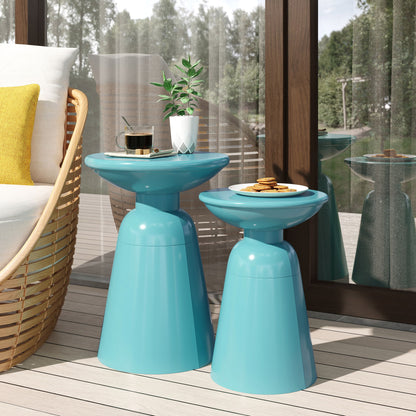 Two Serenita Outdoor Side Tables With Flared Design (Set of 2) in blue hold a plant, coffee, and cookies on a wooden patio beside a wicker chair with yellow and white pillows, next to glass doors reflecting the scene.