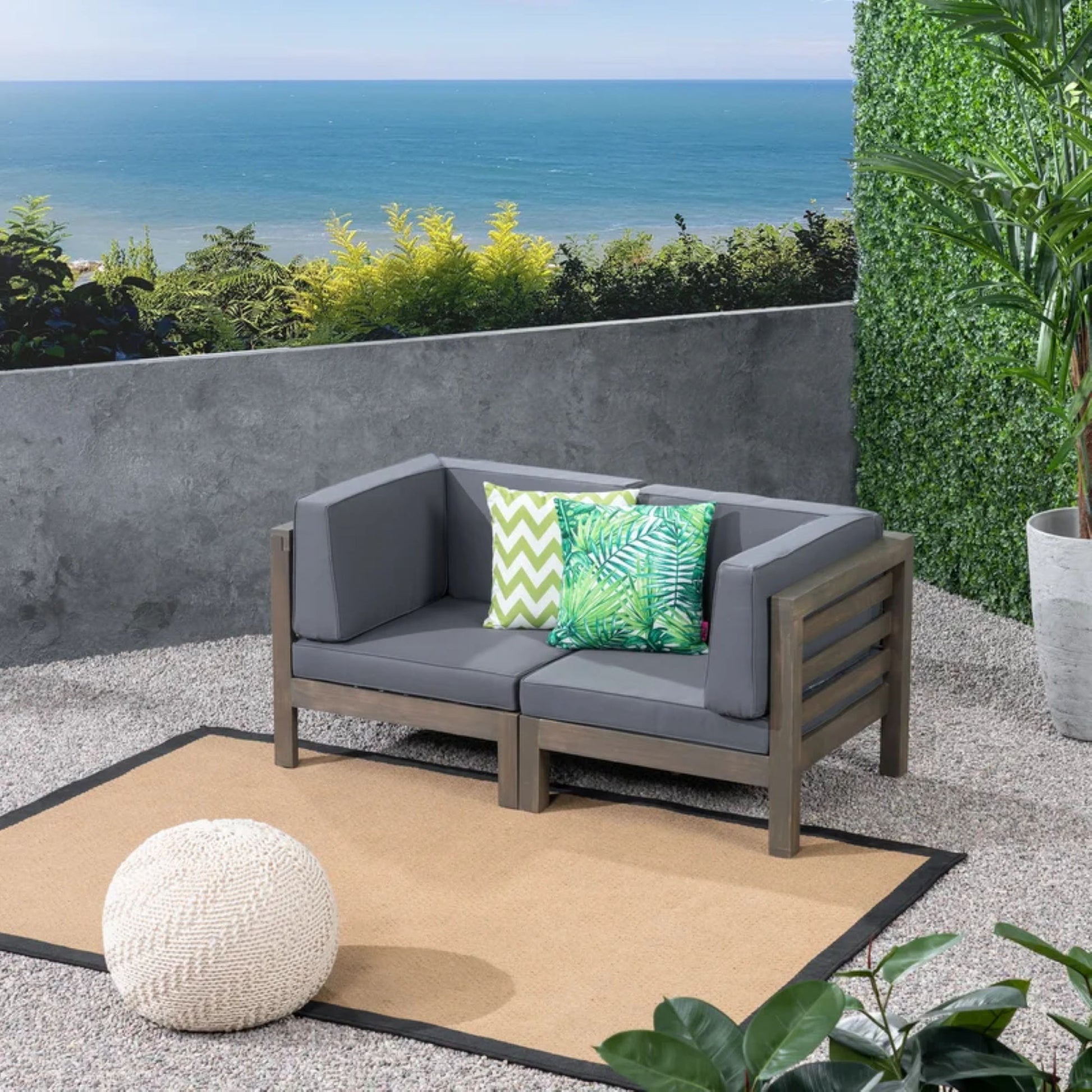 A modern patio with the Oana 2 Piece Loveseat Set featuring gray water-resistant cushions, two patterned green pillows, a tan rug, a round knitted pouf, potted plants, and an ocean view beyond a concrete wall and lush greenery.