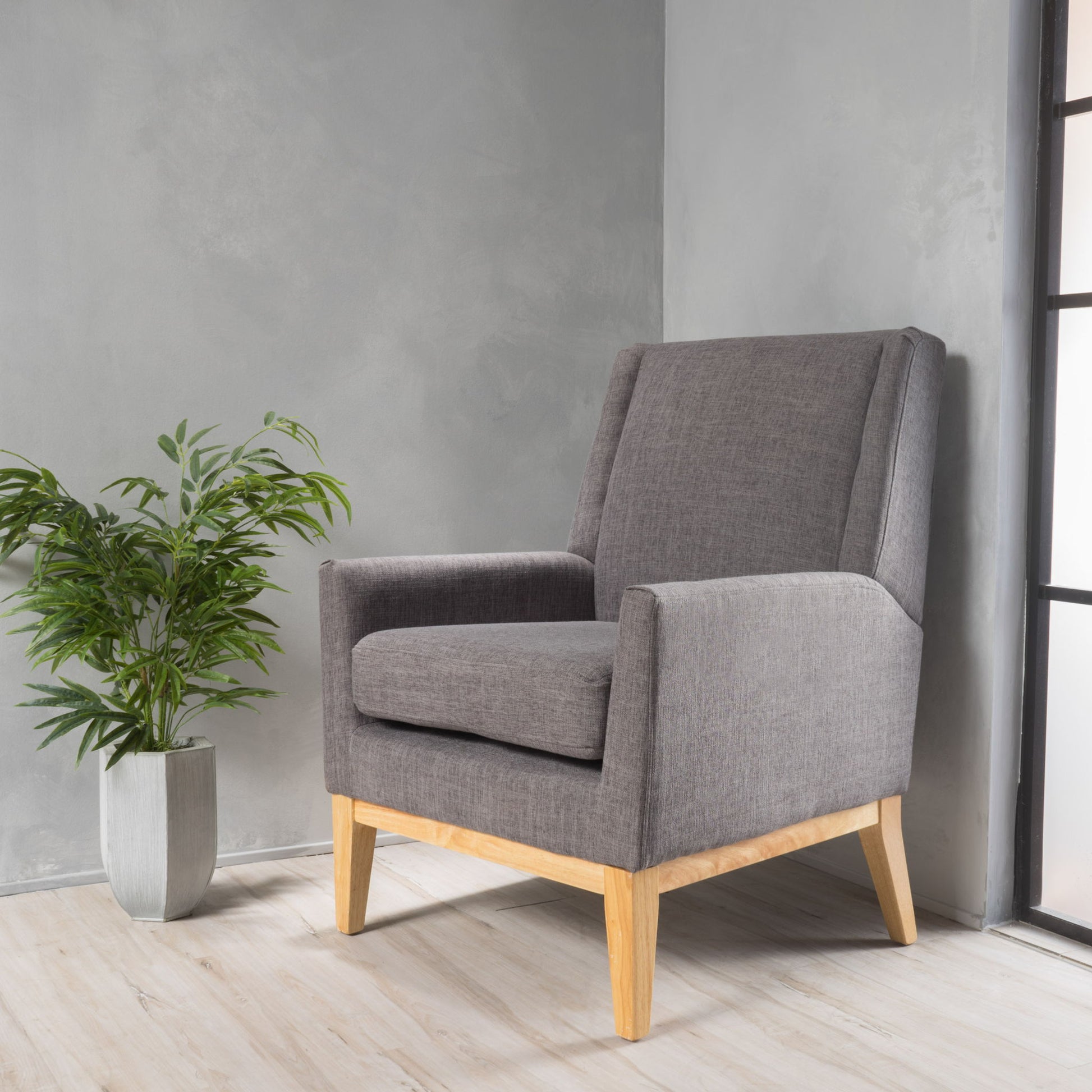 The Modern Accent Chair With Padded Cushion Seat, featuring gray upholstery and wooden legs, is placed on a light wood floor beside a green potted plant, set against a gray textured wall and a black-framed window.