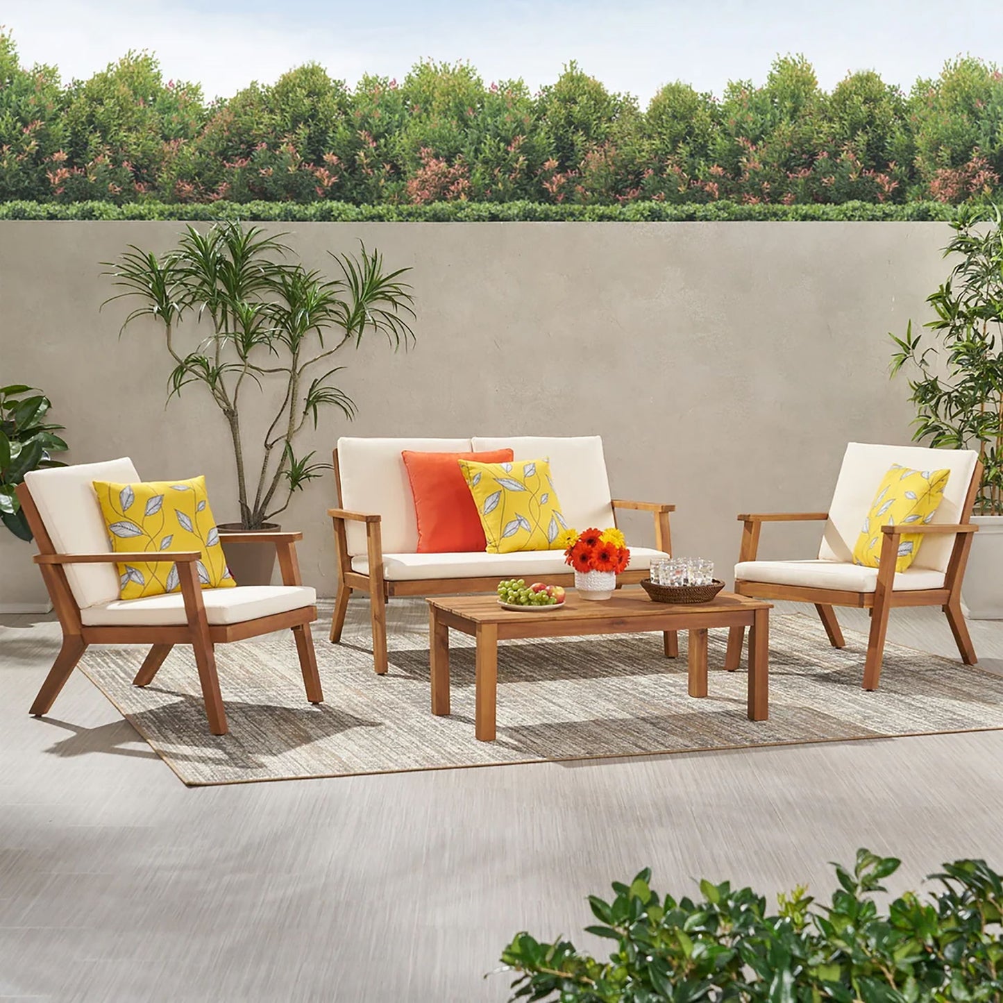 The Avacyn Outdoor Acacia Wood 4 Seater Chat Set features wooden furniture with cream cushions, colorful pillows, a rectangular coffee table, and decorative plants on a rug, set before a concrete wall and trees.