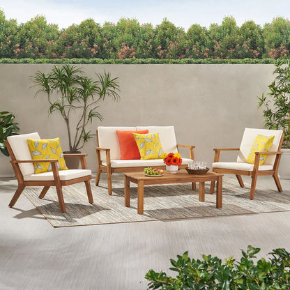 The Avacyn Outdoor Acacia Wood 4 Seater Chat Set features wooden furniture with cream cushions, colorful pillows, a rectangular coffee table, and decorative plants on a rug, set before a concrete wall and trees.