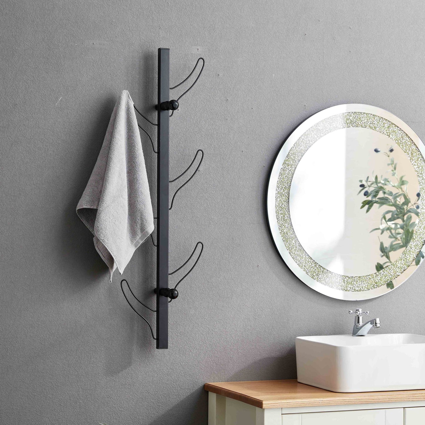 A Vertical Eight Hook Coat Rack Wall Mount in black holds a gray towel next to a round mirror above a white sink and wood vanity, all set against a gray bathroom wall.