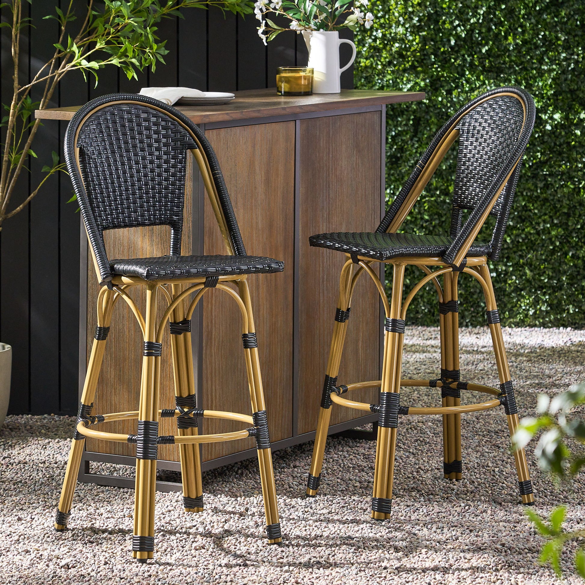Two Outdoor Rattan And Aluminum French Barstools (Set of 2) with black woven seats and backs are placed by a wooden outdoor bar on a gravel patio, surrounded by greenery and potted plants.