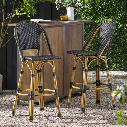 Two Outdoor Rattan And Aluminum French Barstools (Set of 2) with black woven seats and backs are placed by a wooden outdoor bar on a gravel patio, surrounded by greenery and potted plants.