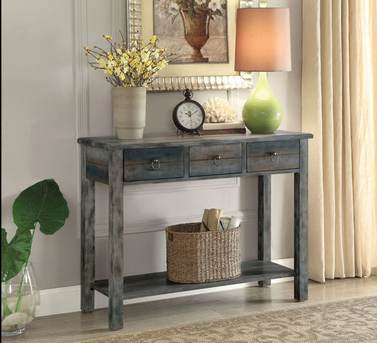 The Glancio Console Table in gray features three drawers holding a green lamp, clock, vase with yellow flowers, coral decor, and books. A woven basket with books rests on the lower shelf. A mirror is mounted behind the table.