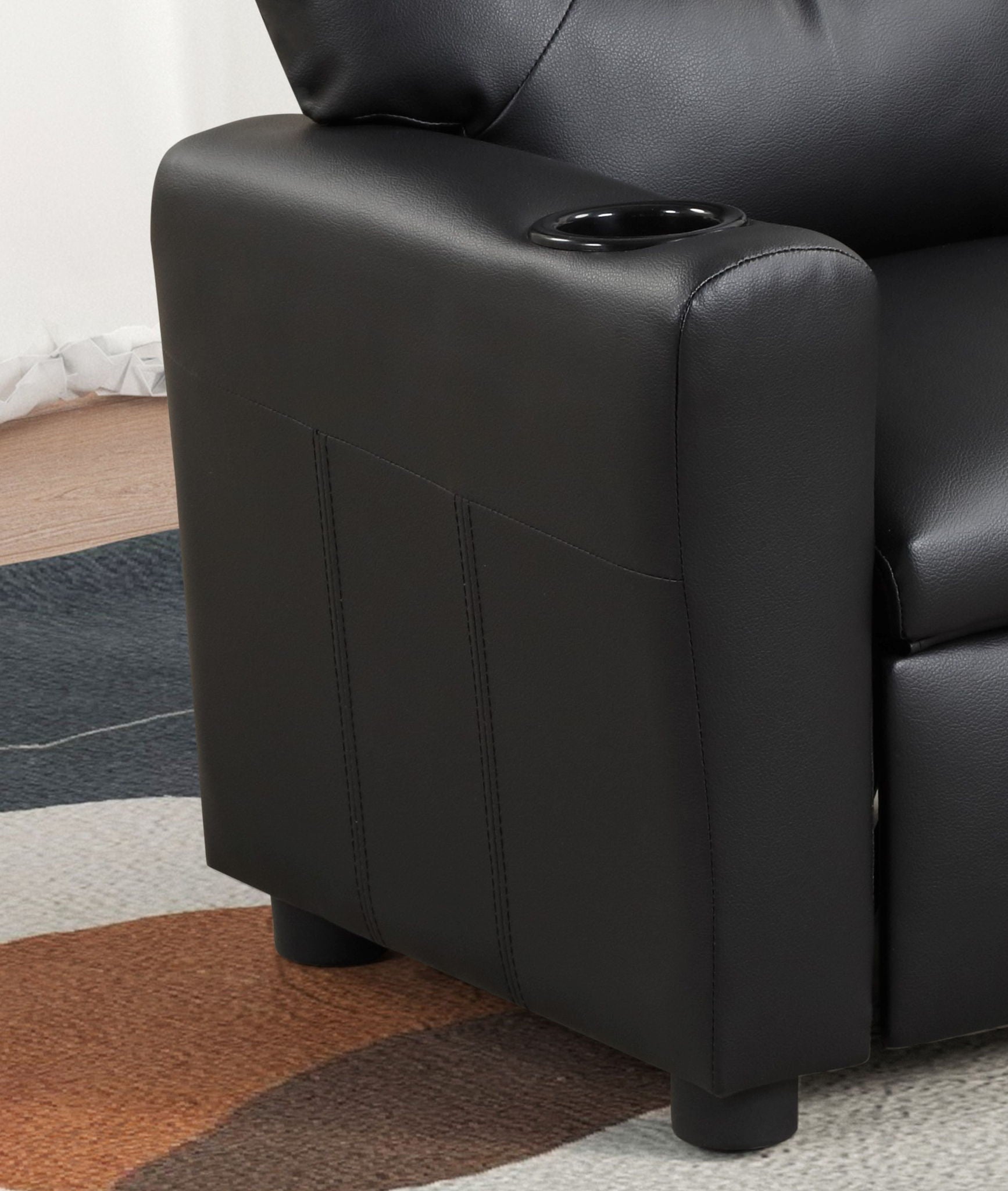 The image shows the armrest of the Marisa - PU Leather Kids Recliner Chair with a built-in cupholder. The chair features short round legs, and part of a colorful patterned rug is visible on the floor.