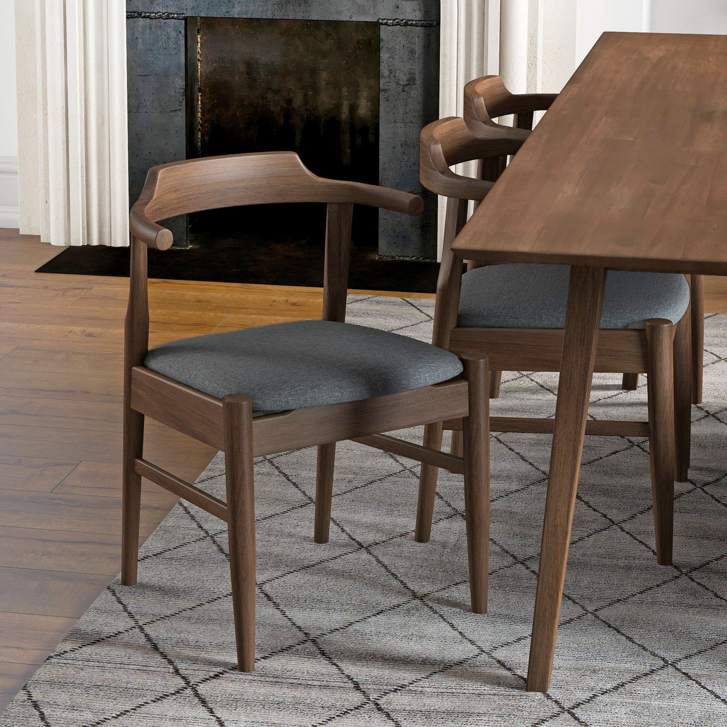 A set of Dakota Mid-Century Modern Velvet Dining Chairs with curved backs and dark gray cushions is placed by a matching dining table atop a patterned gray rug, with wood floors and a fireplace in the background.
