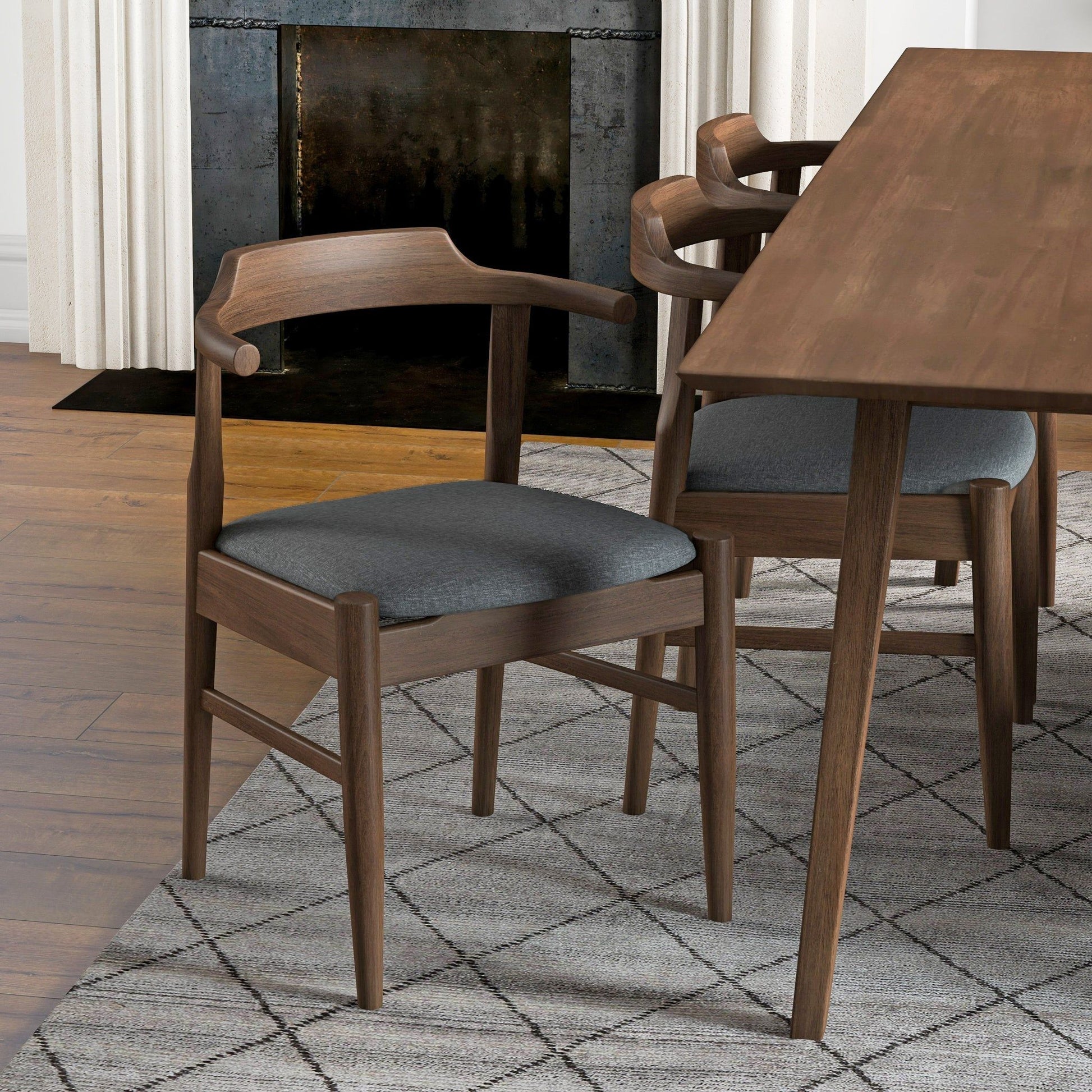 A set of Dakota Mid-Century Modern Velvet Dining Chairs with curved backs and dark gray cushions is placed by a matching dining table atop a patterned gray rug, with wood floors and a fireplace in the background.