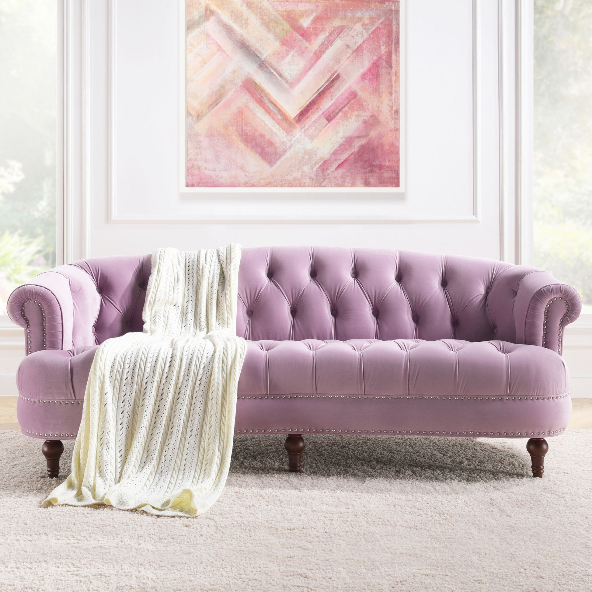 The La Rosa - Victorian Chesterfield Tufted Sofa, in light purple with wooden legs and silver nailhead trim, is draped with a cream knitted throw and placed on a beige rug beneath a pink abstract painting in a bright room.