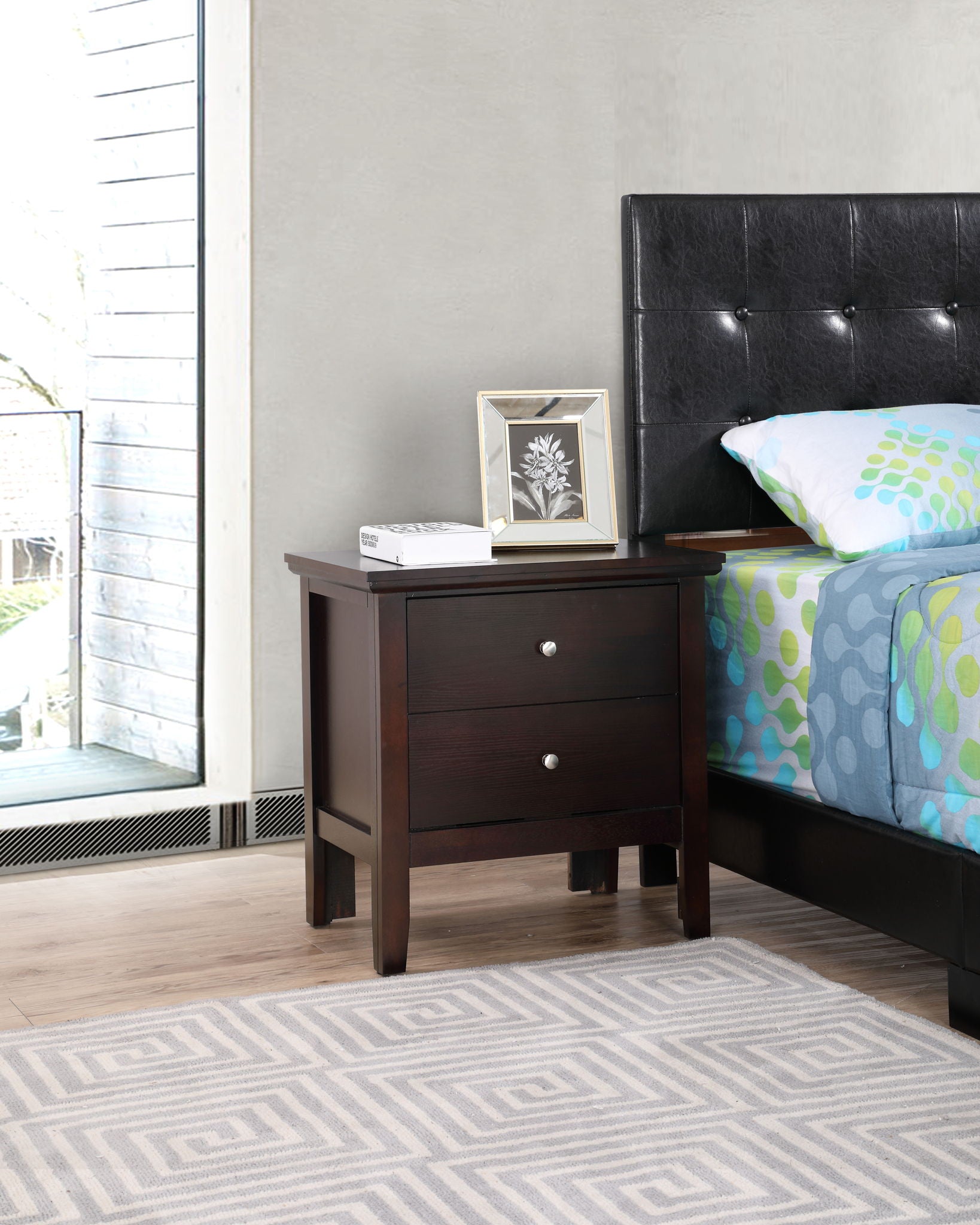 The Primo - Nightstand, featuring a dark wood finish, two drawers with silver knobs, stands by a bed with a blue and green patterned duvet. A framed photo and book rest atop it on a light-toned geometric rug.