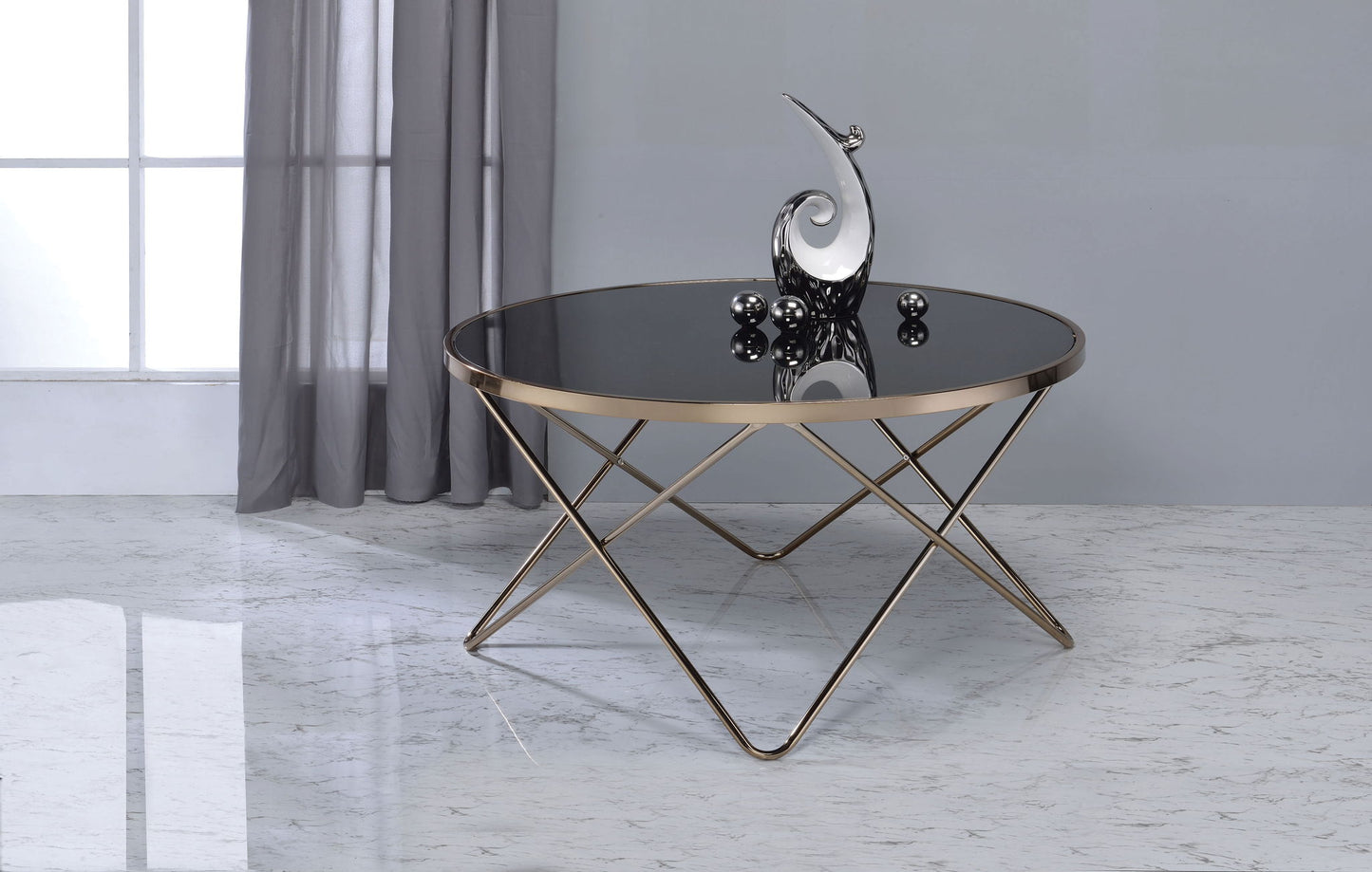 The Valora Glass Coffee Table with gold metal legs sits on a marble floor, topped with a modern silver sculpture featuring decorative spheres. Sheer gray curtains and a large window form the background.