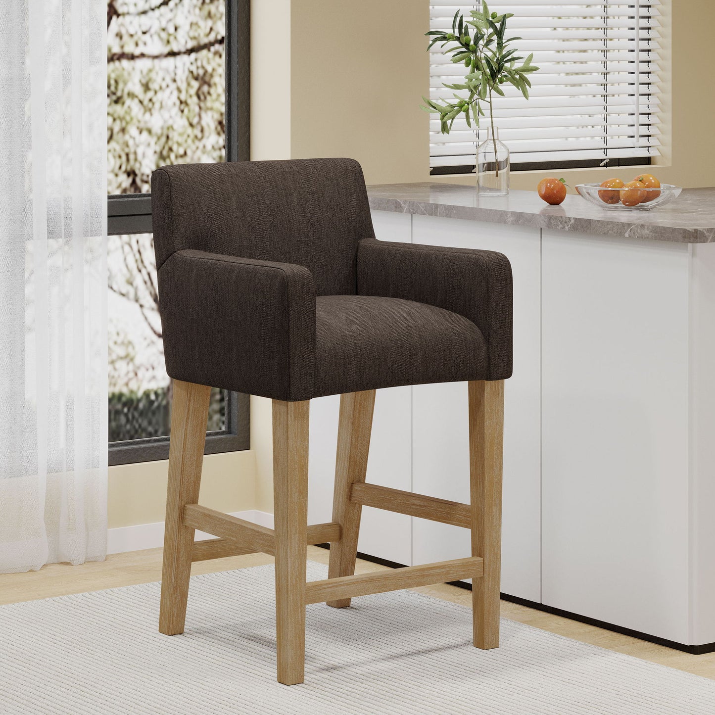The Charmed Upholstered Counter Stool With H Frame Base And Round Arms, in dark brown with wooden legs, sits on a light rug in a modern kitchen near a bright window, alongside a counter topped with a small plant and oranges.
