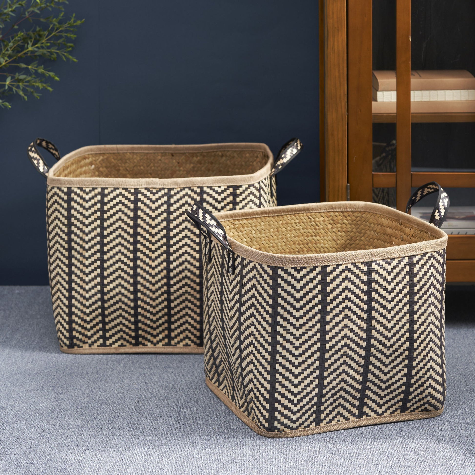 A set of two square palm leaf woven wicker storage baskets with black and brown zigzag patterns and handles are placed on a gray carpeted floor beside a wooden cabinet.