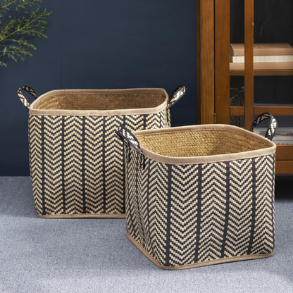 A set of two square palm leaf woven wicker storage baskets with black and brown zigzag patterns and handles are placed on a gray carpeted floor beside a wooden cabinet.
