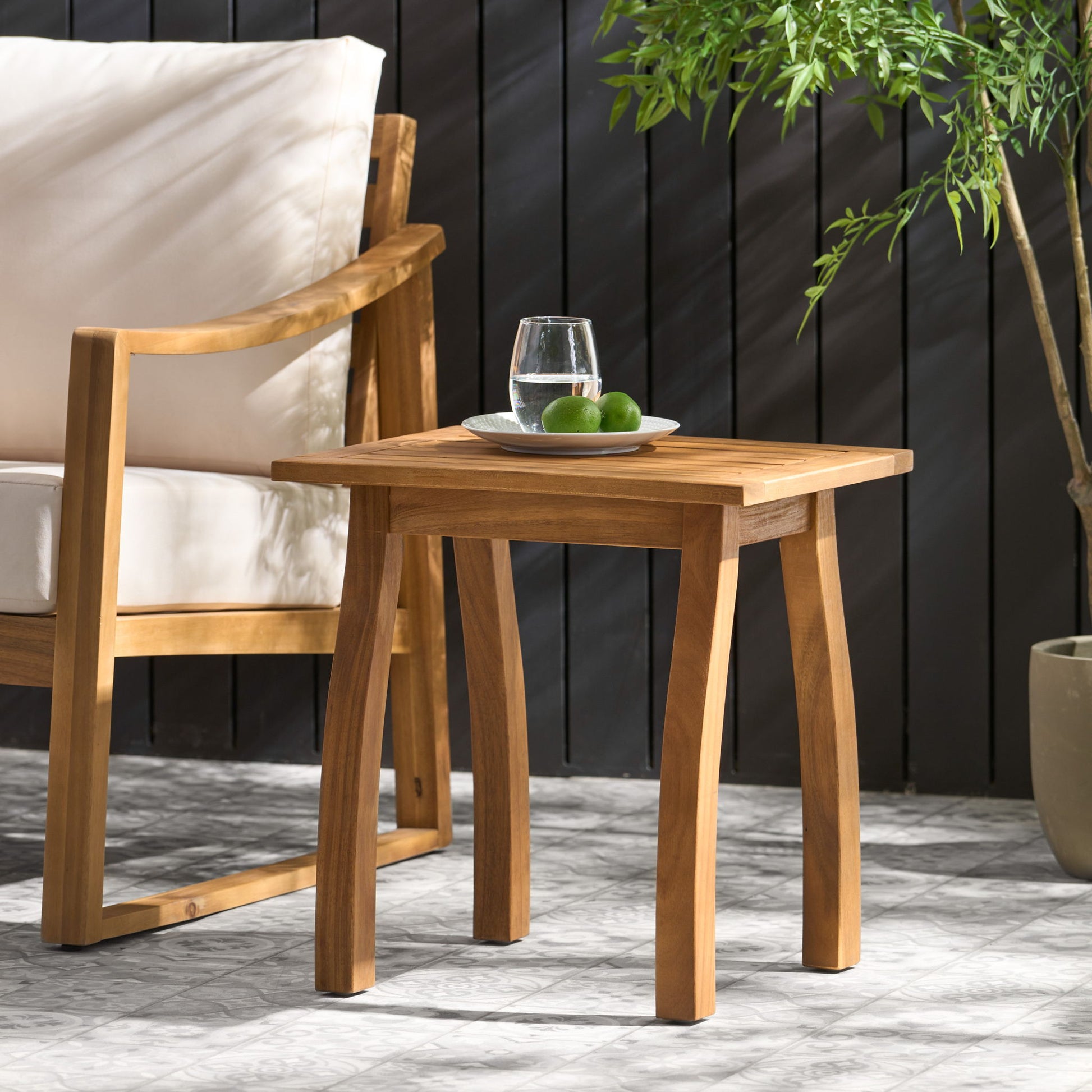 The Kinesis Outdoor Side Table with a slatted top and wood frame holds a glass of water and two limes beside a cushioned wooden chair, with a potted plant nearby and a dark wooden fence in the background.