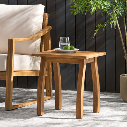 The Kinesis Outdoor Side Table with a slatted top and wood frame holds a glass of water and two limes beside a cushioned wooden chair, with a potted plant nearby and a dark wooden fence in the background.