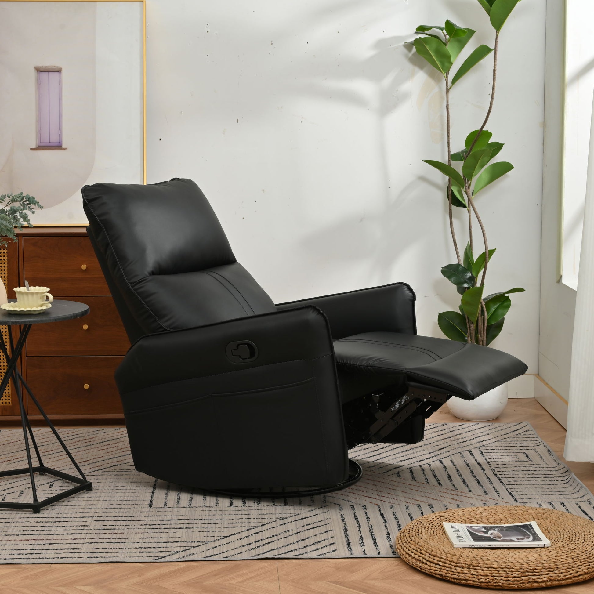 A 360° Modern Small Rocking Swivel Recliner Chair With Side Pocket, in black with an extended footrest, sits on a gray patterned rug beside a small round table, a green potted plant, and a woven mat holding a magazine in a bright room.