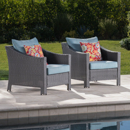 Two Antibes Armchairs with water-resistant blue cushions and red floral throw pillows sit side by side on a stone poolside patio, bordered by a green lawn and shrubs.