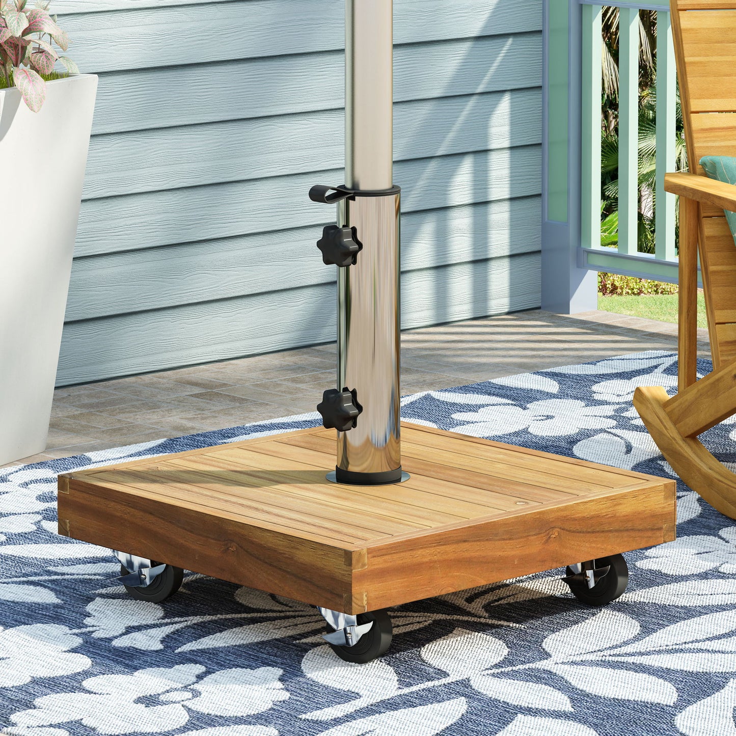 A close-up of the Ralph - Square Umbrella Base in teak with wheels supporting a metal pole, set on a blue and white floral outdoor rug by a wooden rocking chair on a porch.