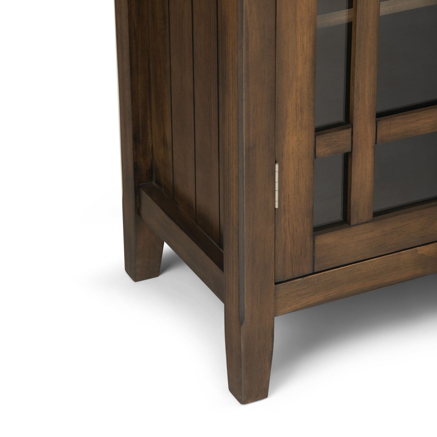Close-up of the bottom corner of the Bedford - Storage Media Cabinet with a medium brown finish, highlighting its legs, side panel, and part of the glass-paneled door frame.