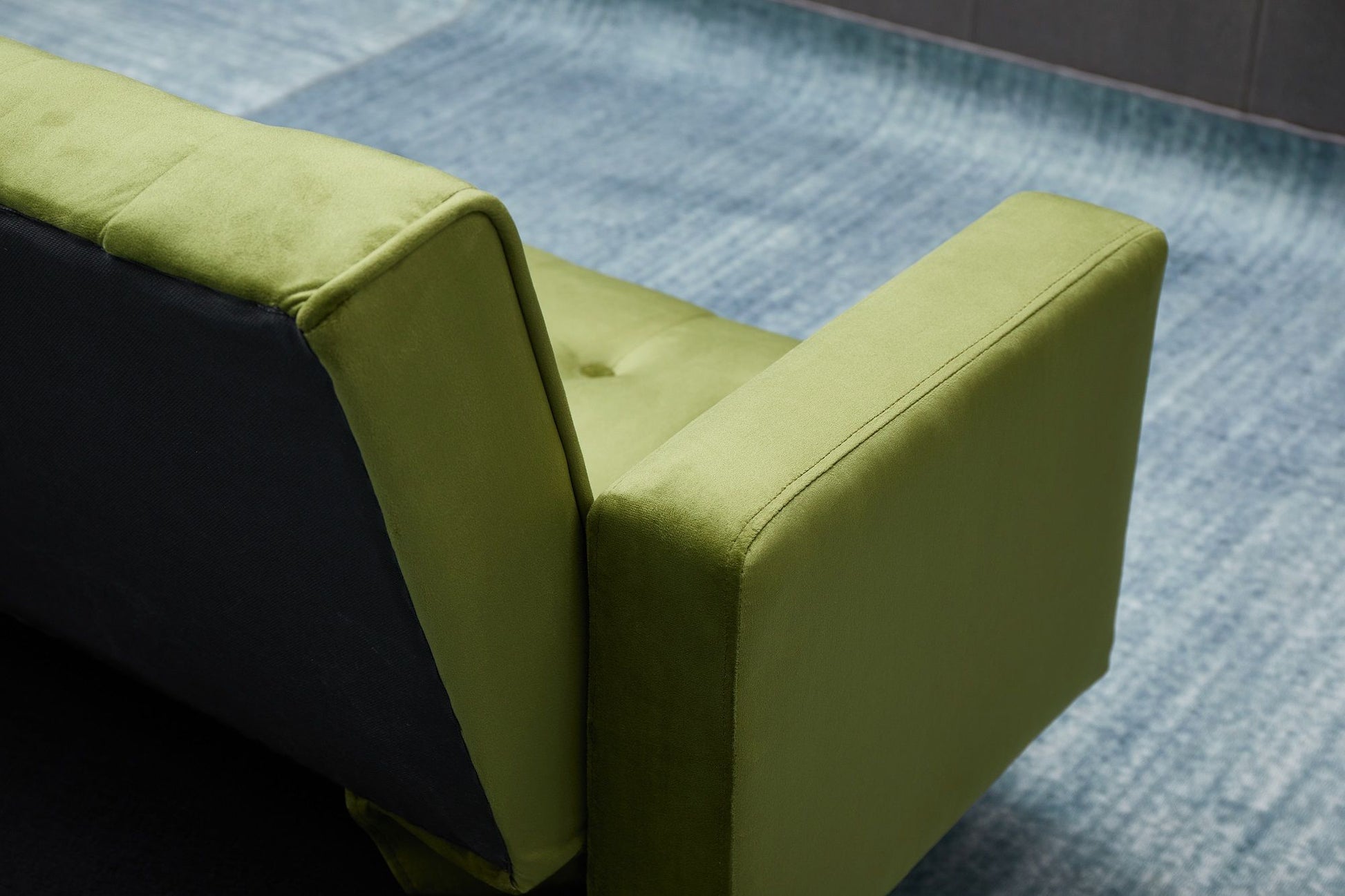 A close-up of the Comfort Compact Design Sofa Bed, featuring green velvet upholstery on its back and armrest, set atop a blue textured carpet.