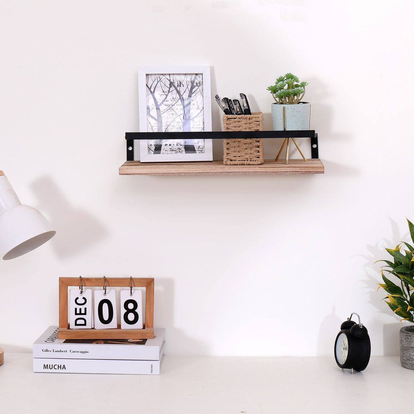 A minimalist desk setup features a calendar showing December 8, lamp, black clock, and plant. Above, natural wood floating wall shelves hold a framed photo, plant, and two woven containers with pens.