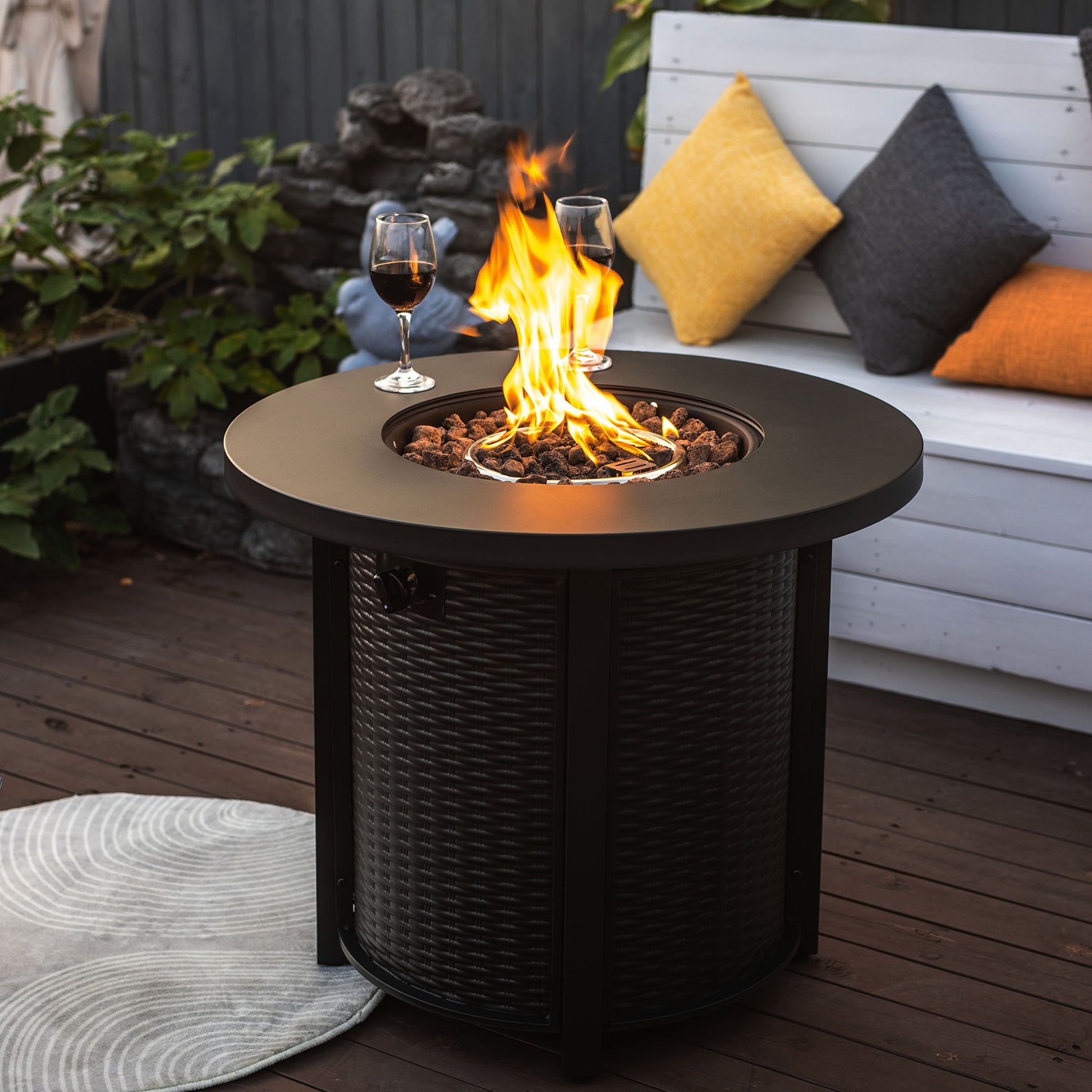 A 30" Propane Round Fire Table (40000Btu, black) with a central flame sits on a wooden deck. Two wine glasses rest atop it. A white bench with yellow and gray pillows and nearby garden plants complete the cozy setting.
