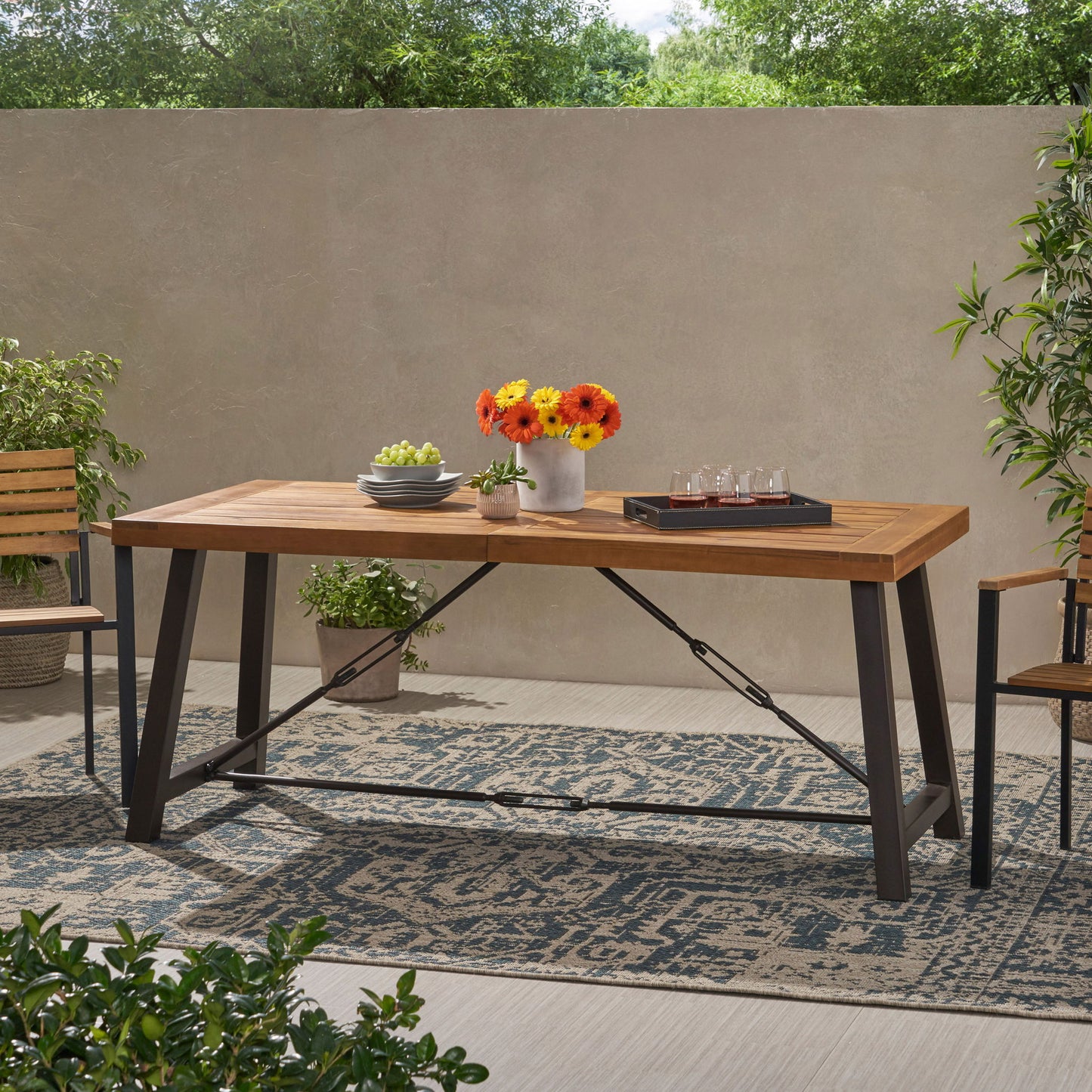 The Outdoor Dining Table With Slat Wooden Panel And Turnbuckle Accents - Teak sits on a patterned rug, topped with flowers, a small potted plant, and a tray. Surrounded by wooden chairs, greenery, and a beige wall.