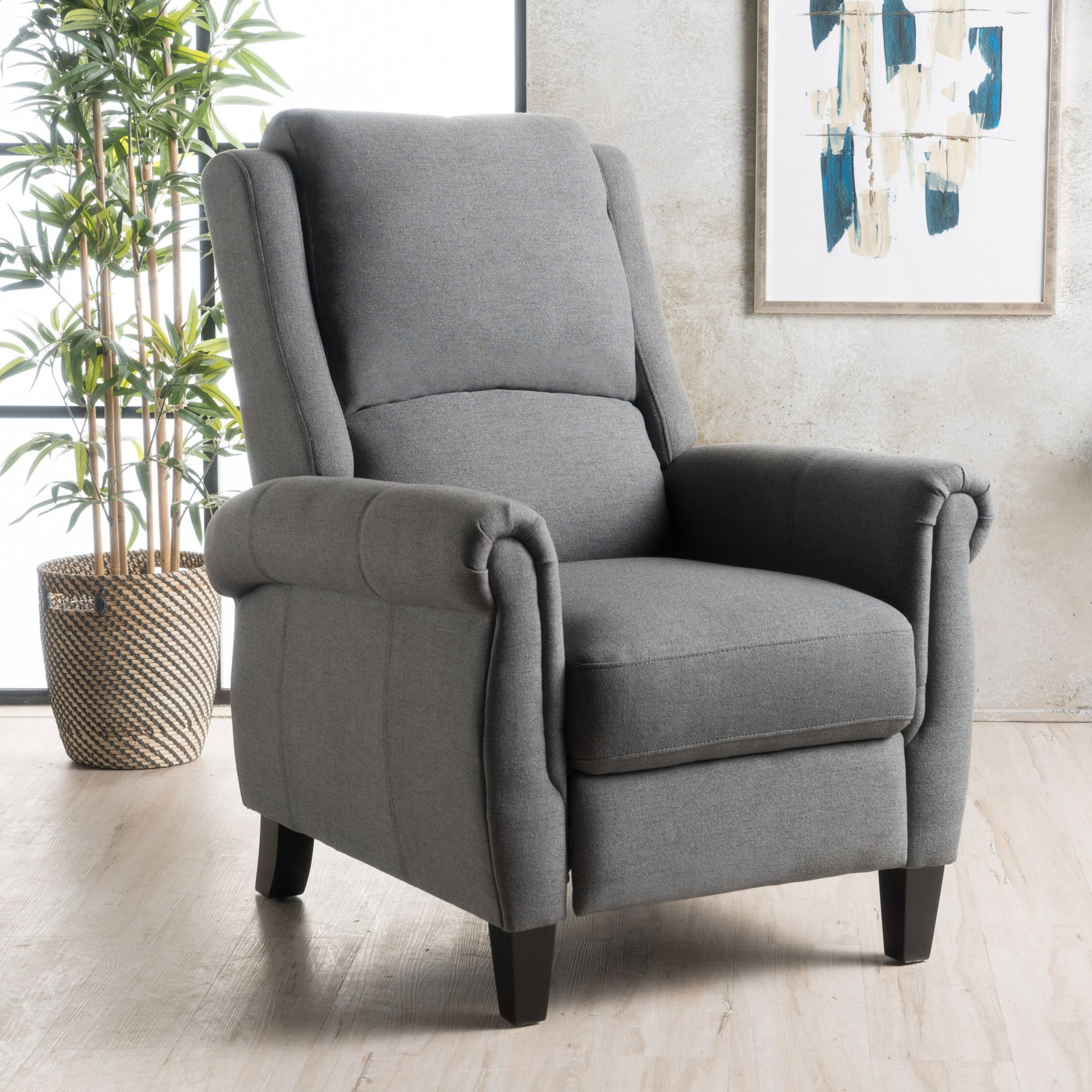A Contemporary Push Back Recliner Chair with gray upholstery and dark wood legs is positioned on a light wood floor beside a potted plant, with abstract art displayed on the wall behind it.