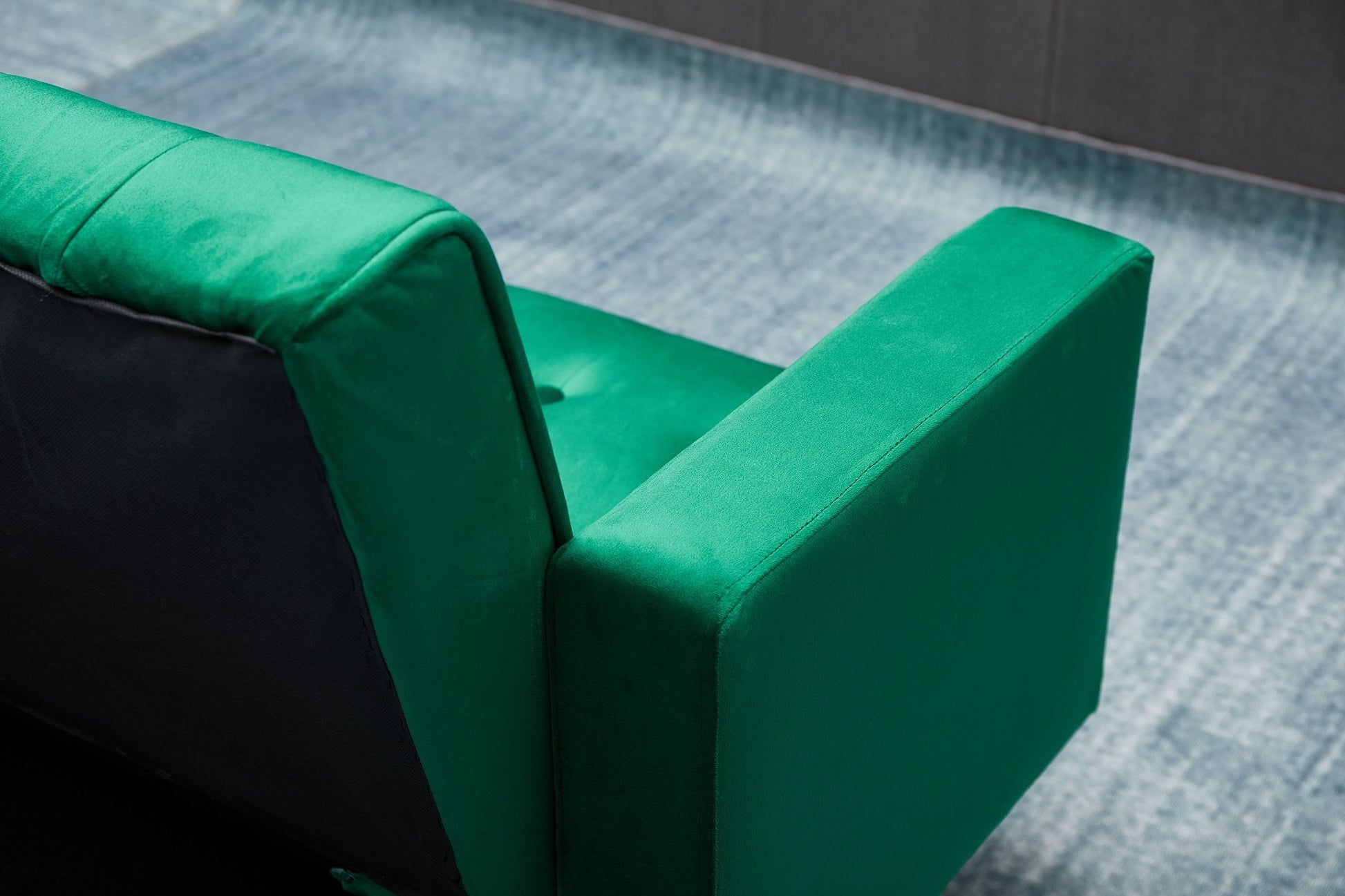 A close-up of the back and armrest of the Comfort Compact Design Sofa Bed in green velvet, set on a light blue textured carpet with a dark wall as the backdrop.