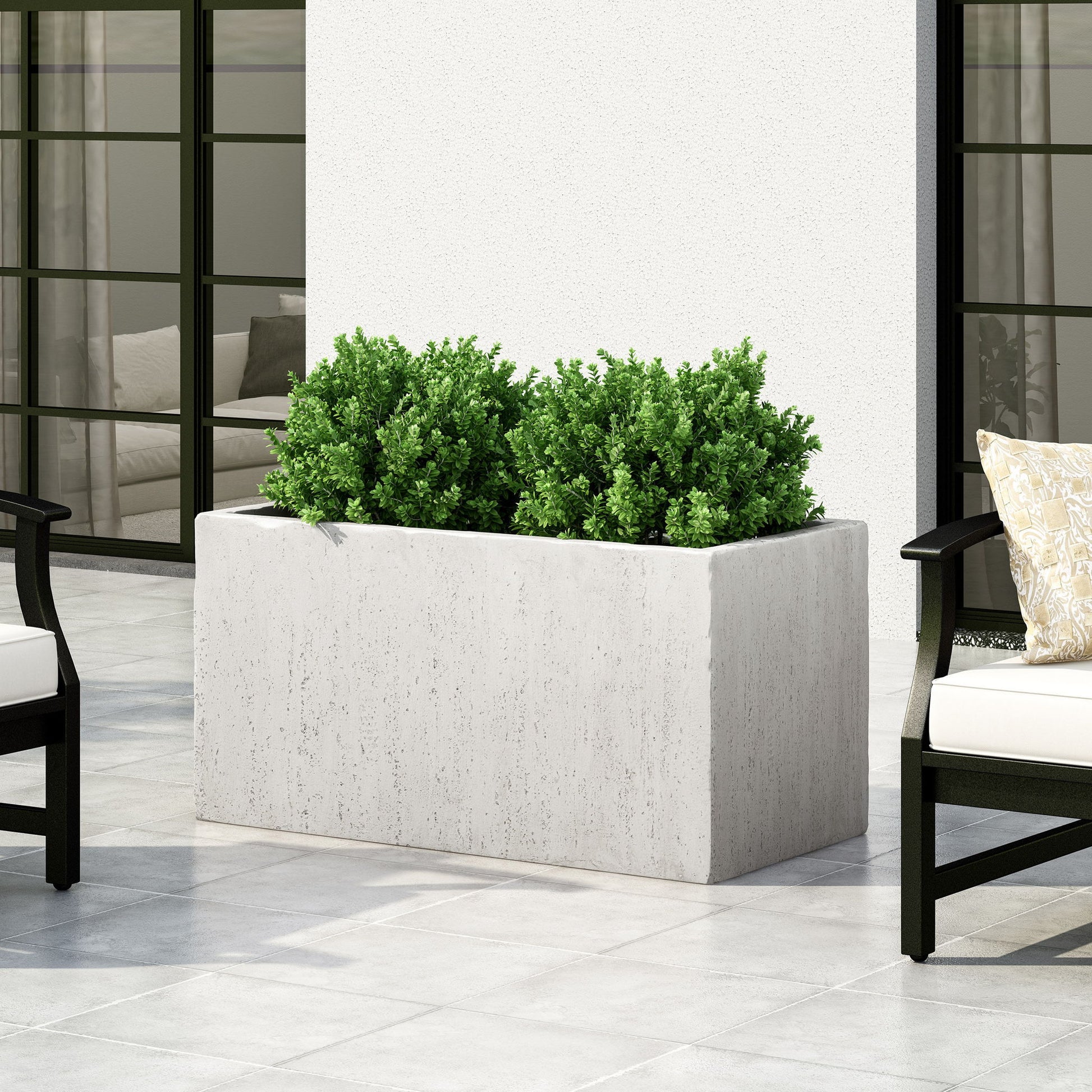 The Modern Outdoor Planter, a rectangular white concrete piece with green shrubs, rests on a light gray tiled patio near black-framed chairs with white cushions, with large windows and an indoor seating area in the background.