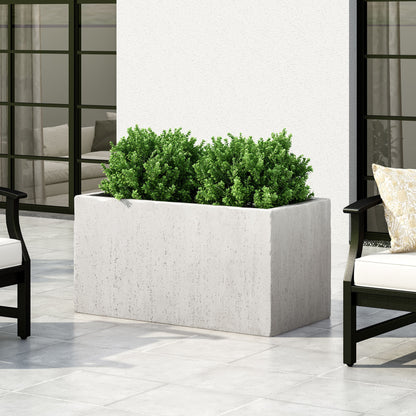 The Modern Outdoor Planter, a rectangular white concrete piece with green shrubs, rests on a light gray tiled patio near black-framed chairs with white cushions, with large windows and an indoor seating area in the background.