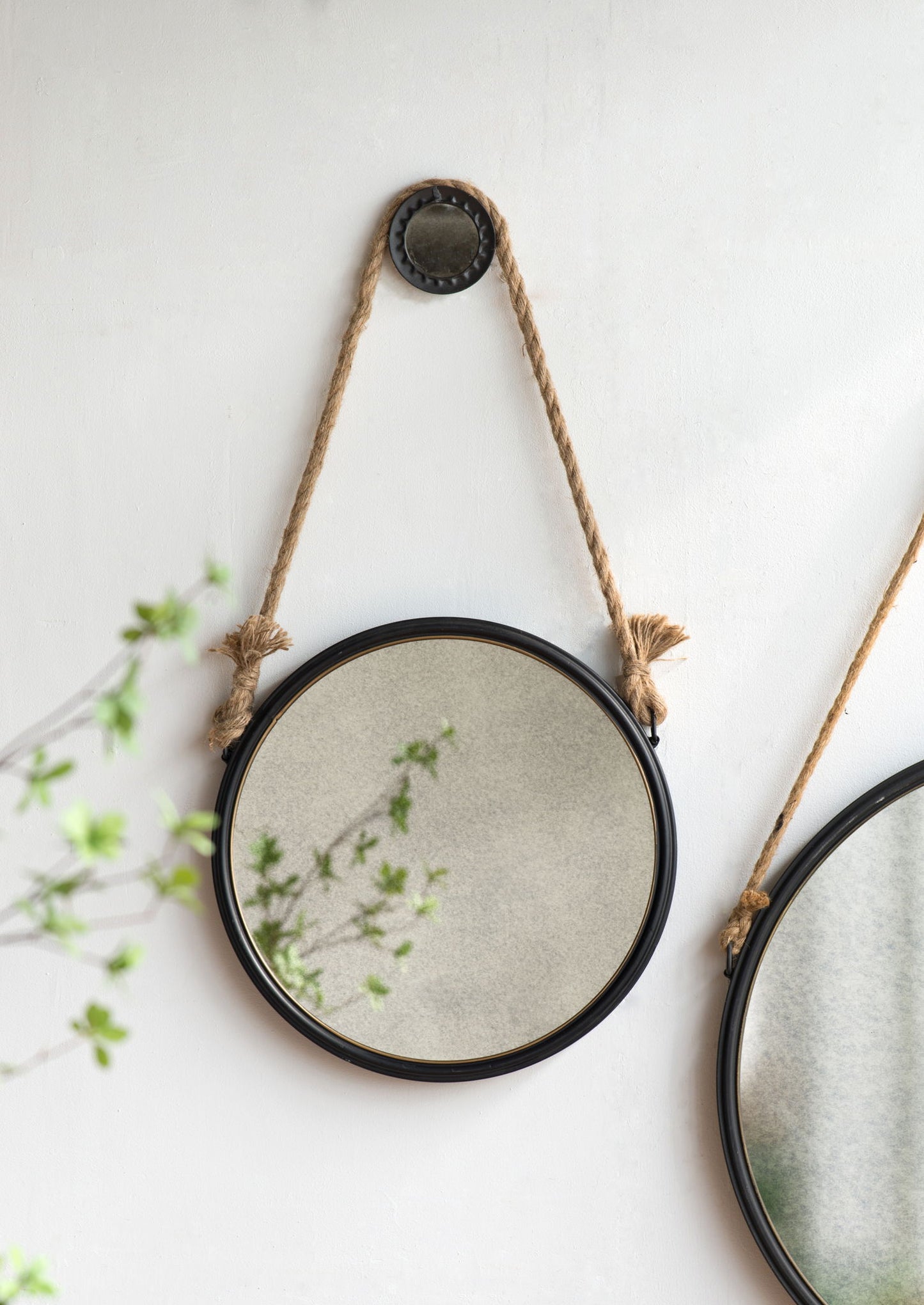 The Handsome Cleveland Mirror With Rope Strap, featuring a contemporary round metal black frame, hangs on a white wall, reflecting soft green branches; another mirror appears partially on the right.