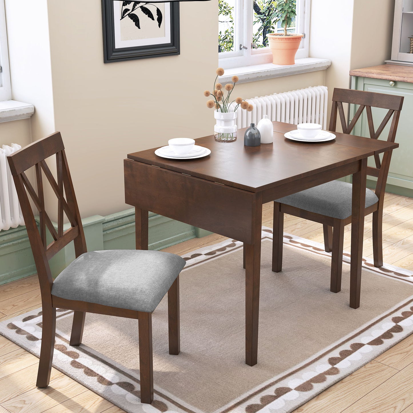 A bright room features the 3 Piece Kitchen Dining Set with Drop Leaf Table and 2 Upholstered Chairs in brown, placed on a beige rug. The table is set with plates and cups, decorative items at the center, with a window and framed art in view.