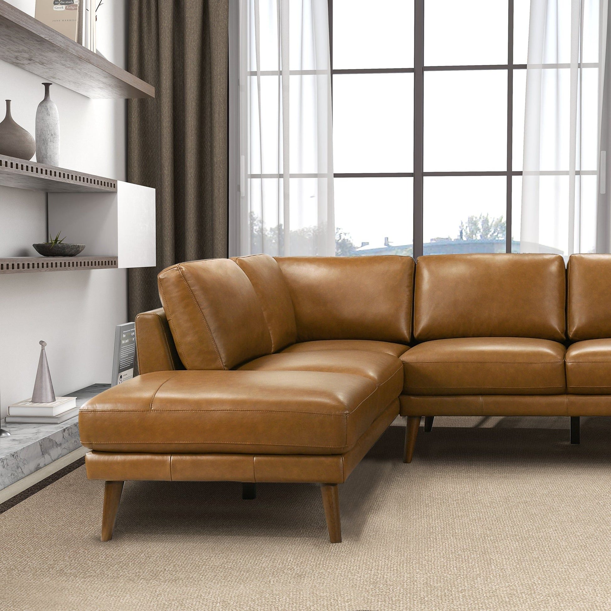 A modern living room featuring the Benson Sectional Sofa With Chaise in tan leather on a beige carpet, with a large window framed by sheer curtains and shelves on the left displaying vases and books.