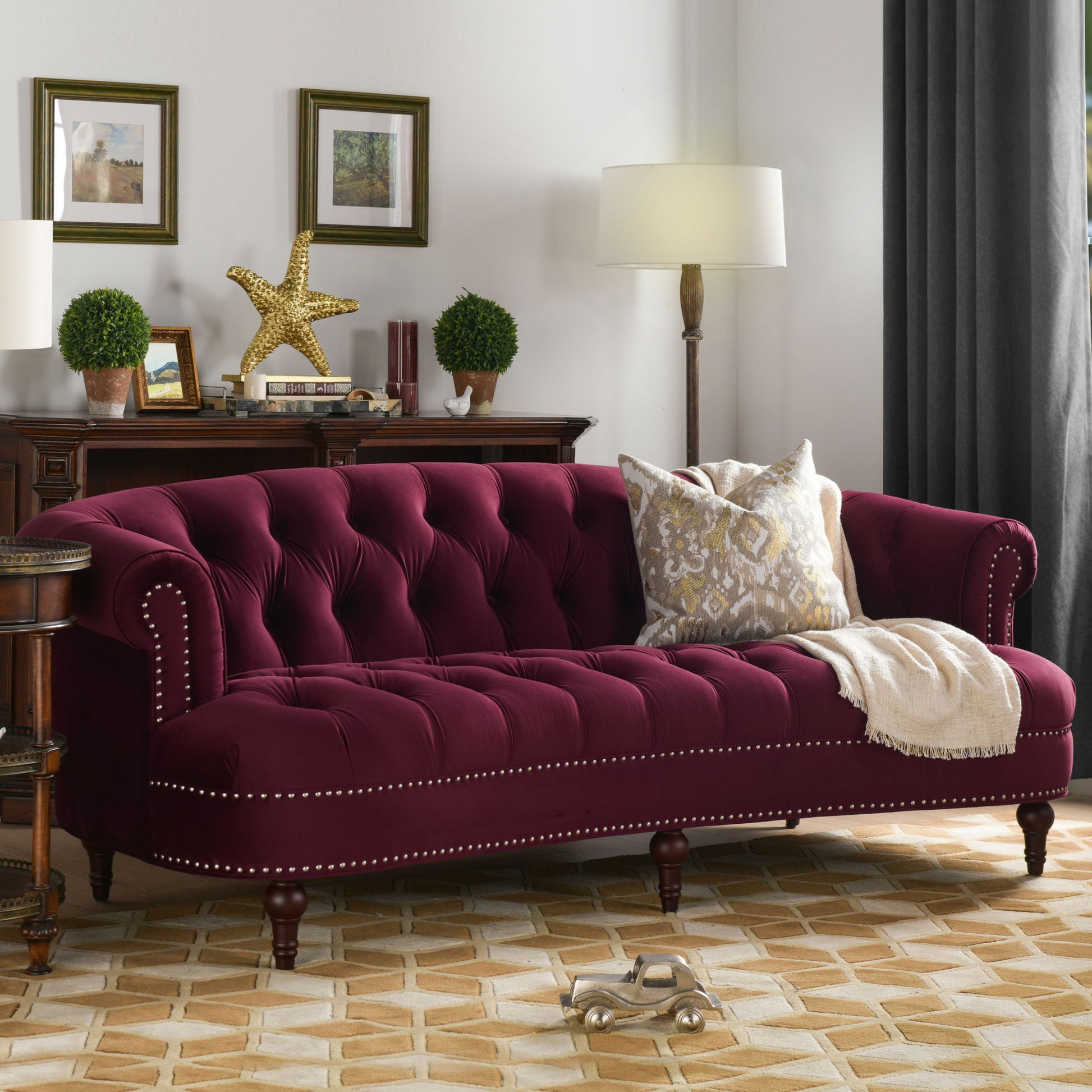A living room features the La Rosa - Victorian Chesterfield Tufted Sofa in burgundy with studded trim, patterned pillows, and a beige throw. Behind it is a dark wood sideboard with decor, two lamps, framed art, and gray curtains over the window.