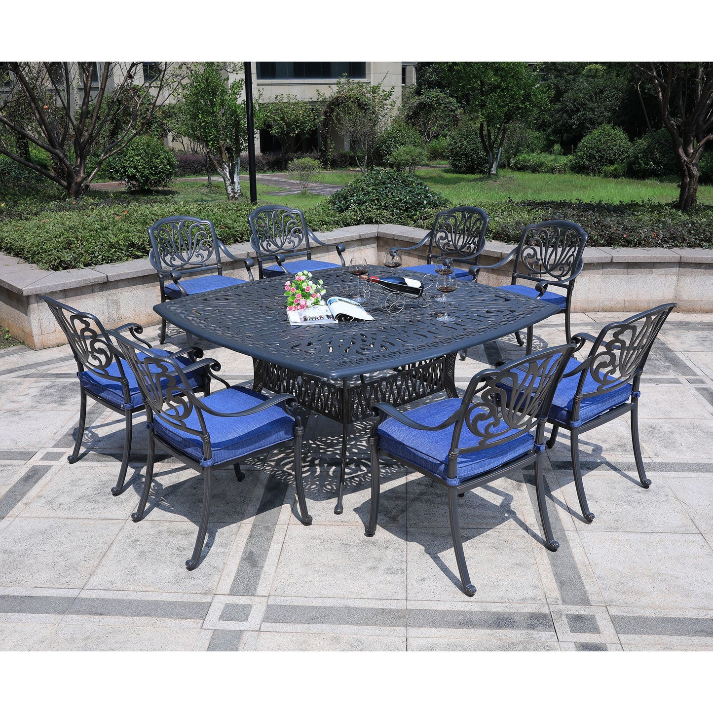The Square 8 Person 64" Long Aluminum Dining Set With Cushions features a black metal square table and eight matching chairs with blue cushions, arranged on a stone patio in a garden setting with greenery.
