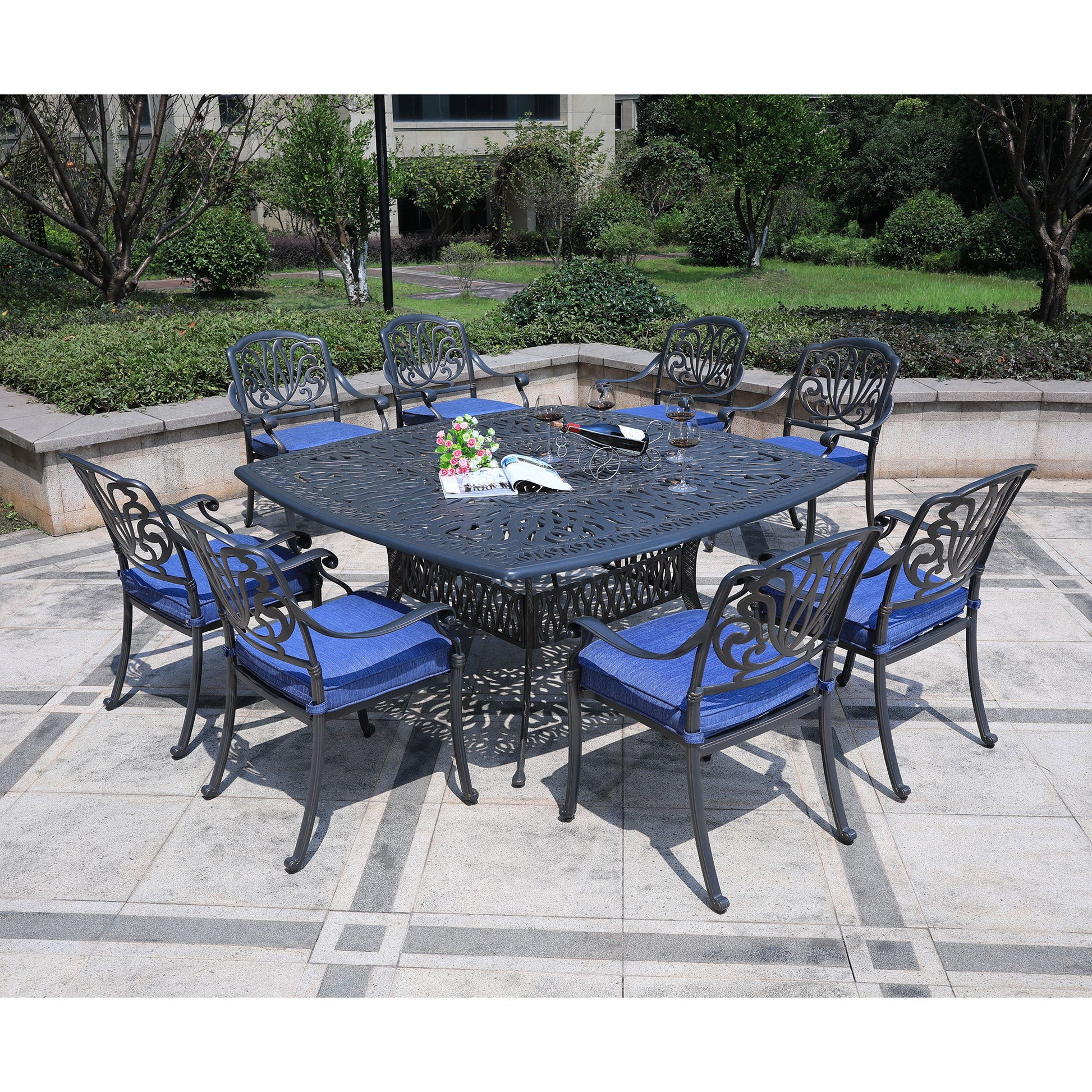 The Square 8 Person 64" Long Aluminum Dining Set With Cushions features a black metal square table and eight matching chairs with blue cushions, arranged on a stone patio in a garden setting with greenery.