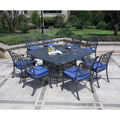 The Square 8 Person 64" Long Aluminum Dining Set With Cushions features a black metal square table and eight matching chairs with blue cushions, arranged on a stone patio in a garden setting with greenery.