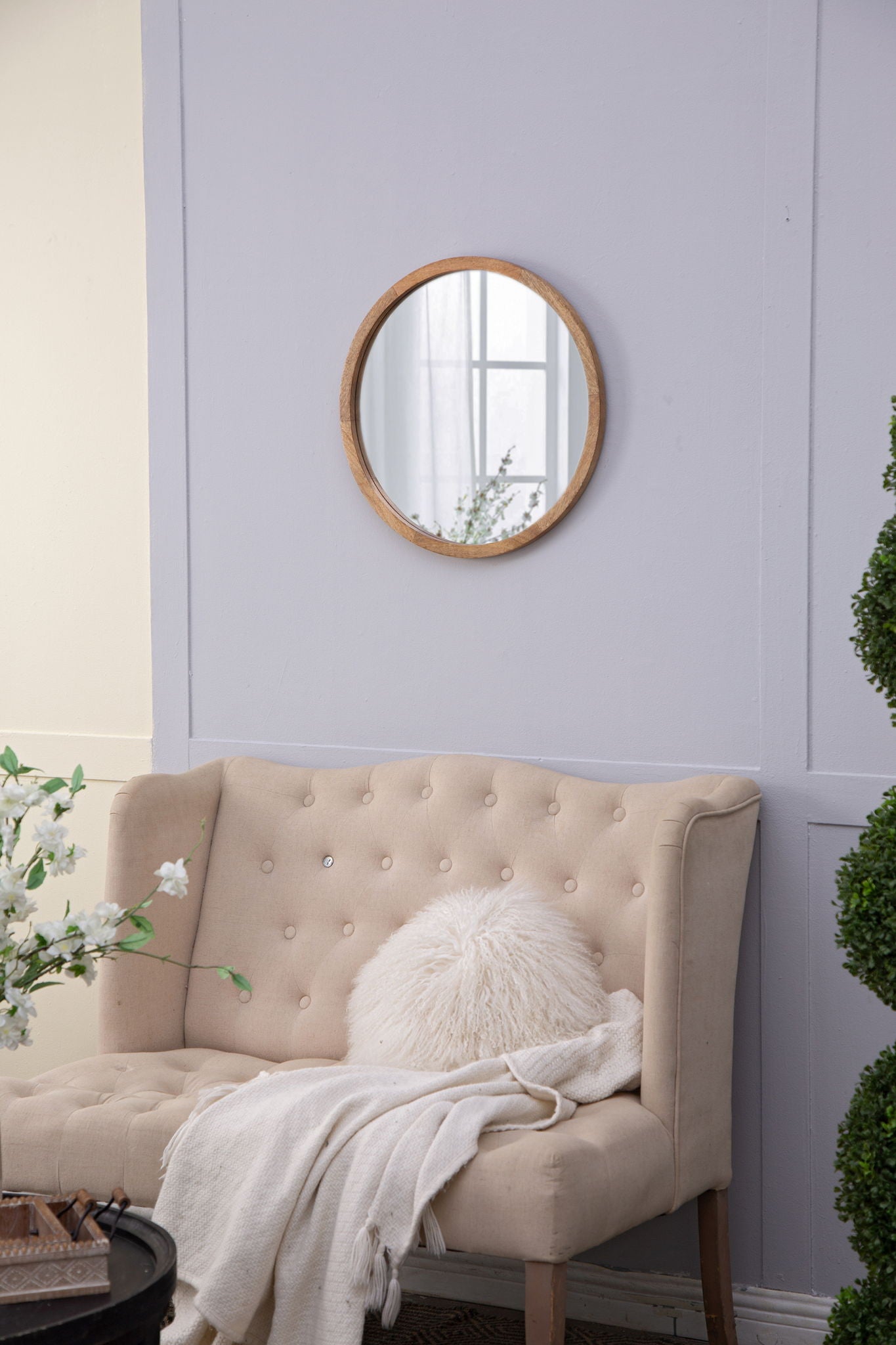 A cozy beige tufted loveseat with a white fluffy pillow and throw sits against a light gray wall. Above, the Circle Wall Mirror With Wooden Frame, Wall Mirror For Living Room hangs, with green plants and white flowers adding freshness.