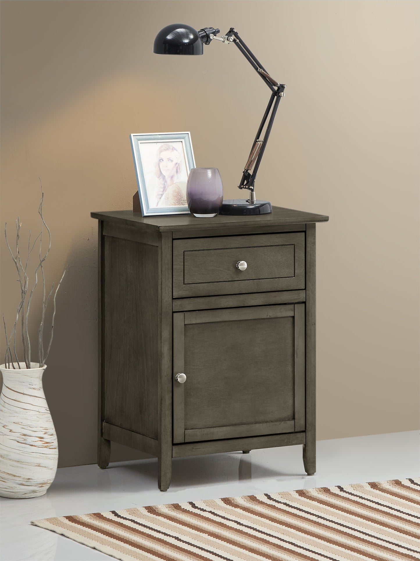 The Izzy 1 Drawer/1 Door Nightstand in gray stands by a beige wall, topped with a black desk lamp, framed photo, and purple vase. A tan vase with twigs sits on the floor nearby, and part of a striped rug can be seen.