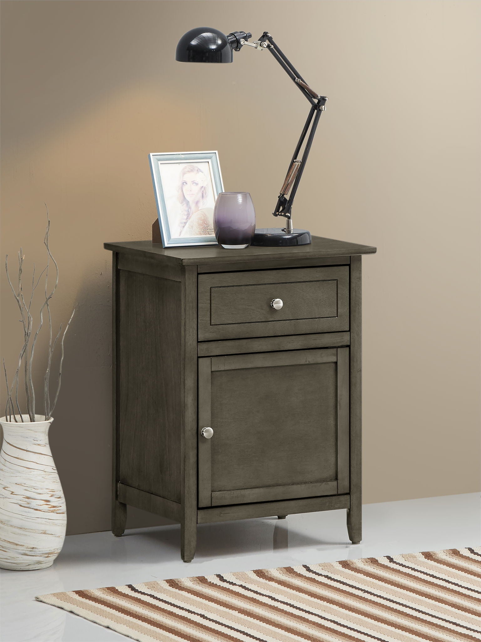 The Izzy 1 Drawer/1 Door Nightstand in gray stands by a beige wall, topped with a black desk lamp, framed photo, and purple vase. A tan vase with twigs sits on the floor nearby, and part of a striped rug can be seen.