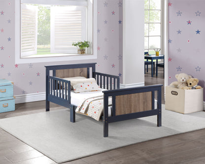 The Connelly Reversible Panel Toddler Bed with blue and wood accents sits on a gray rug in a kids' room with star-patterned wallpaper. Plush toys fill a storage bin, while natural light streams in through the window.