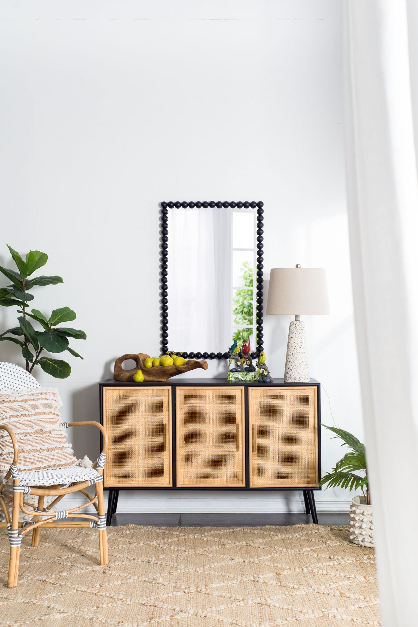 A cozy, modern living space with a rattan chair, woven rug, and wooden cabinet topped with a lamp, fruit bowl, and decor. The Rectangle Mirror, Wall Mirror For Living Room Bedroom Entryway - Black hangs above; a tall plant sits nearby.