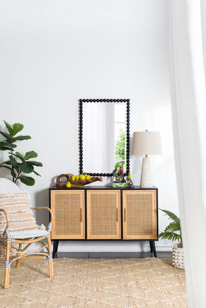 A cozy, modern living space with a rattan chair, woven rug, and wooden cabinet topped with a lamp, fruit bowl, and decor. The Rectangle Mirror, Wall Mirror For Living Room Bedroom Entryway - Black hangs above; a tall plant sits nearby.