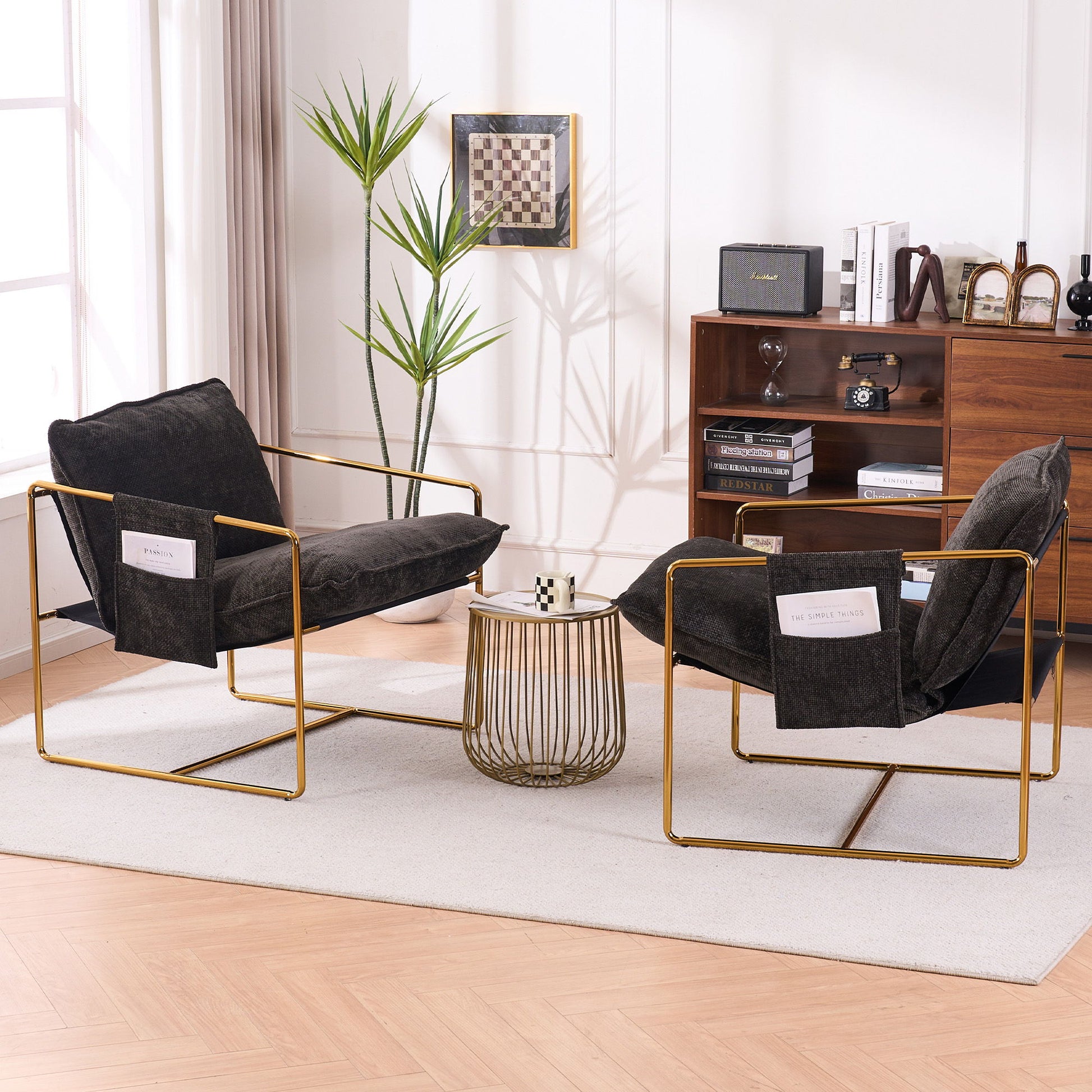 Two Upholstered Hanging Armchairs with gold-plated frames, crushed foam cushions, and skin-friendly woven fabric face each other across a round coffee table on a beige rug. A wooden bookshelf, potted plant, and chessboard accent the bright room.