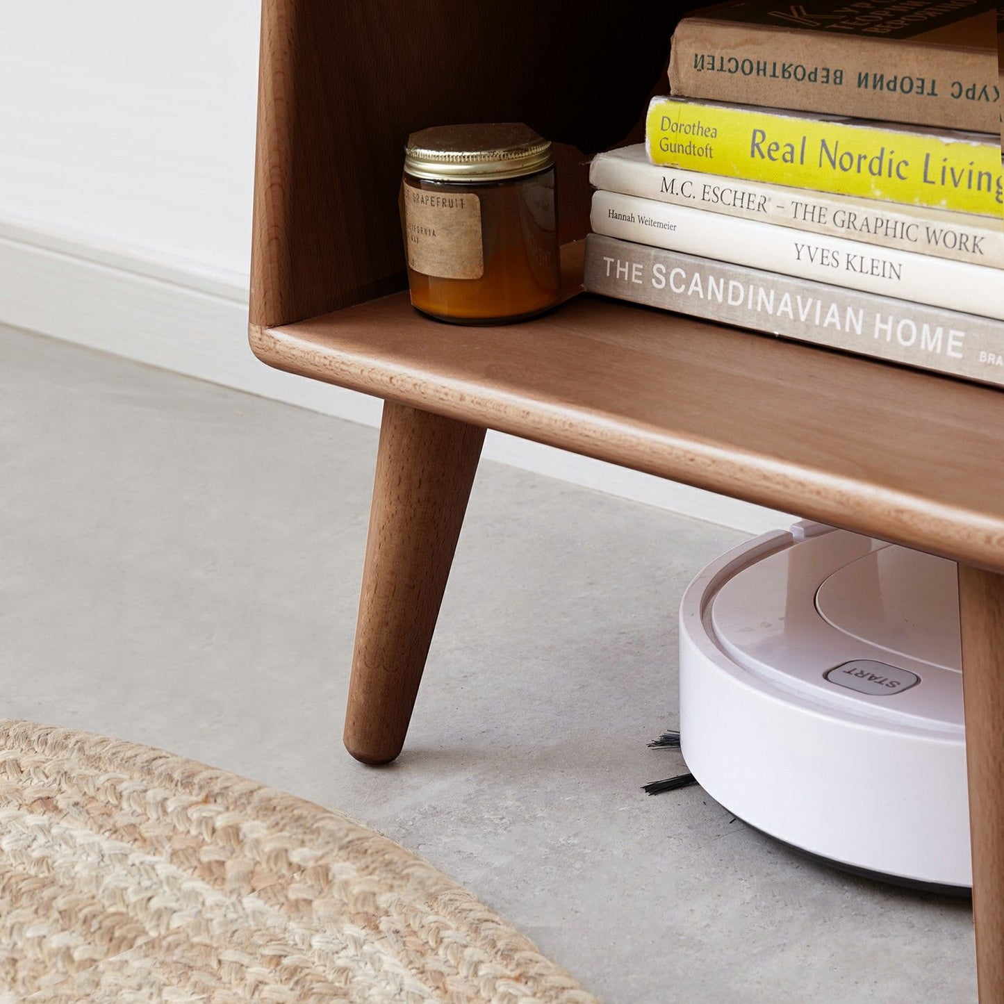 A white robot vacuum cleaner moves under a modern European beech bedside cabinet beside a woven rug on a light floor. Books and a candle jar sit atop the nightstand.