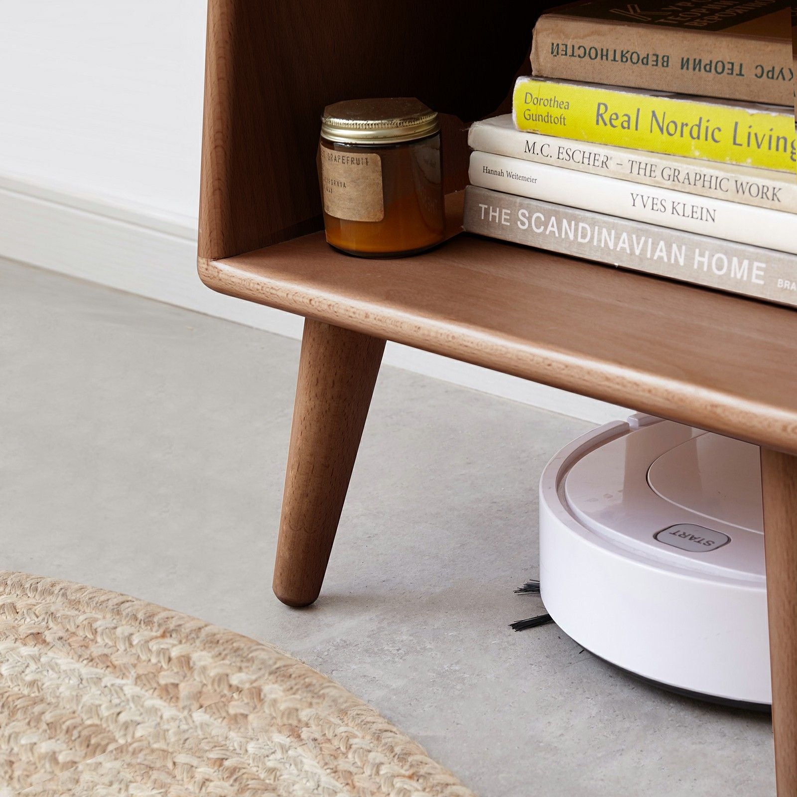 A white robot vacuum cleaner moves under a modern European beech bedside cabinet beside a woven rug on a light floor. Books and a candle jar sit atop the nightstand.