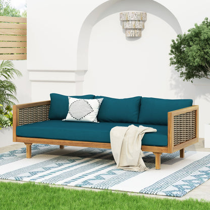 The Claremont 3 Seater Daybed, featuring a teak acacia wood frame and water-resistant teal cushions, sits on a blue and white patterned rug with a cream throw blanket and geometric pillow, framed by plants and a white arched wall.
