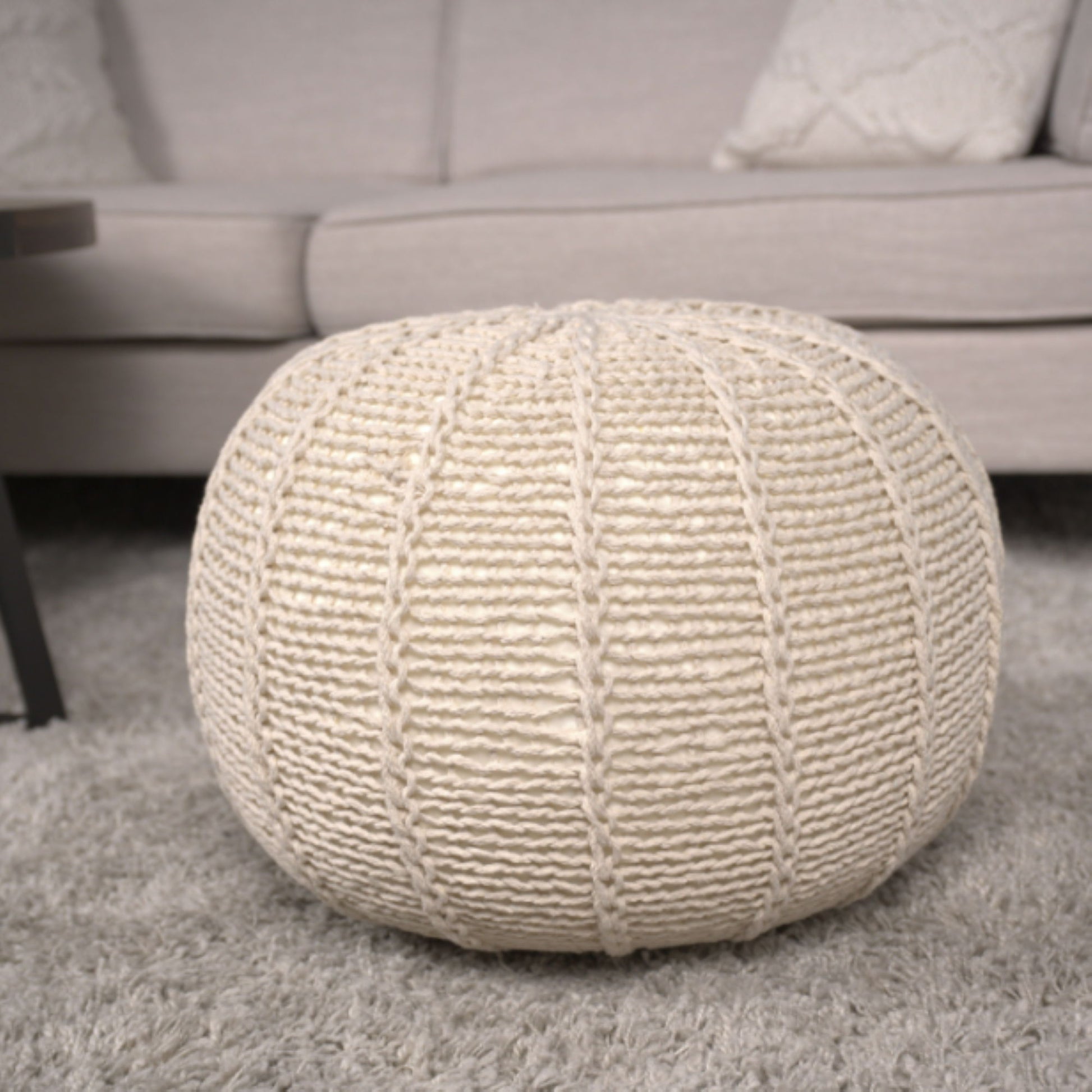 The Bordeaux Knitted Cotton Round Pouf Ottoman and Floor Cushion in cream rests on a light gray shaggy rug before a beige sofa with patterned cushions, creating a cozy living room atmosphere.
